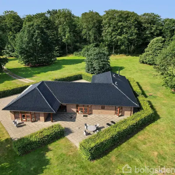 Et moderne hus med sort tegltag omgivet af en flisebelagt terrasse. Huset ligger i en grøn have med tæt hæk og store træer, beliggende nær en skov.