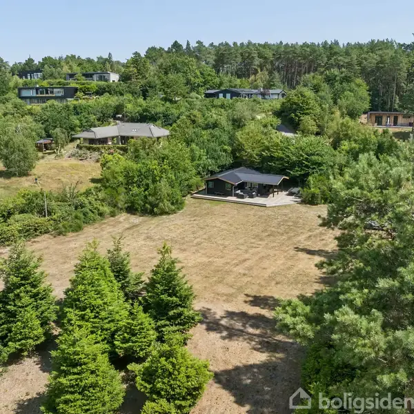 Hus omgivet af grønne træer og buske. Det står på en græsplæne i et kuperet landskab med flere huse spredt i det frodige område under en klar blå himmel.