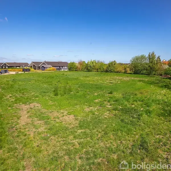 Grønt græsareal strækker sig mod rækkehuse i det fjerne, omkranset af træer under en blå himmel med lette skyer.