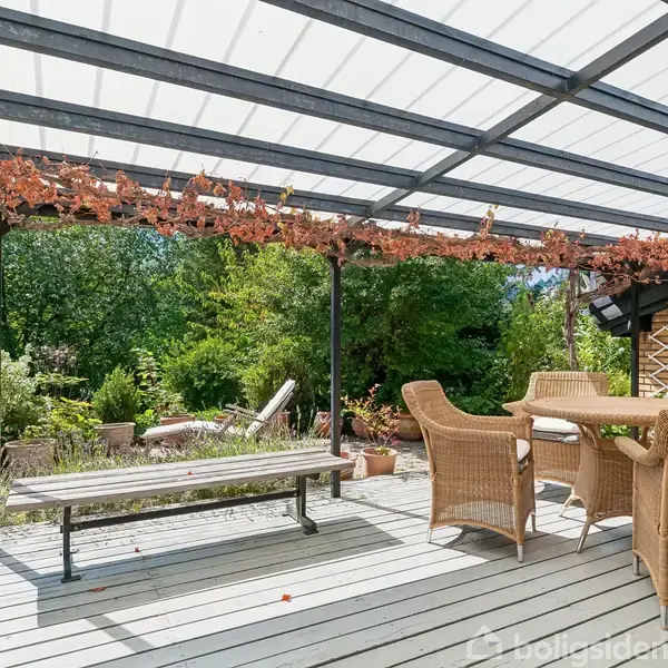 En terrasse med plasttag. Der står en bænk og et sæt med fire fletstole og bord. Baggrunden er en frodig have med træer og buske.