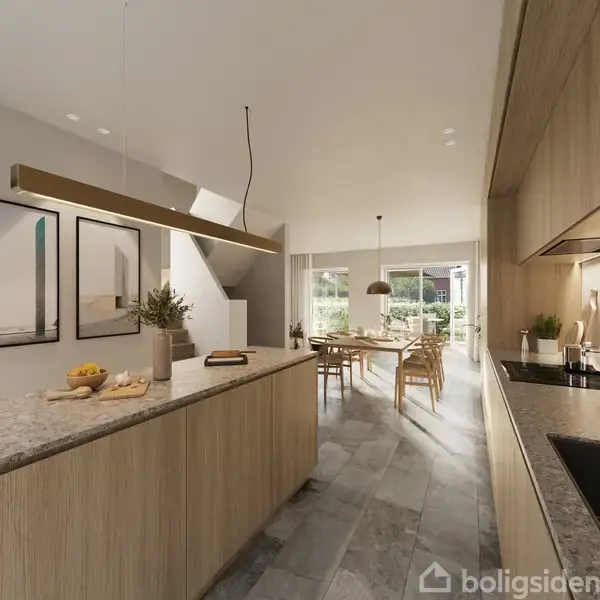 Køkken med træskabe og marmorbordplade, hvorpå der står frugt og en skærebræt. Der er spisebord i baggrunden ved et stort vindue, der lader lys ind.