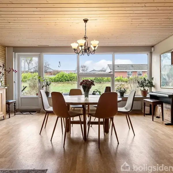 Spisebord står centralt med seks stole, omgivet af træmøbler og store vinduer. Lampe hænger over bordet i lyst rum udsigt til have.