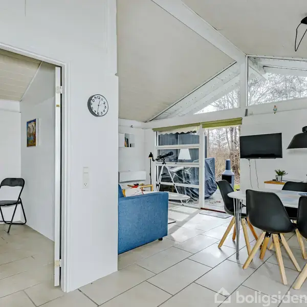 Et moderne, lyst rum med et spisebord omgivet af sorte stole under to pendellamper. I baggrunden ses en blå sofa tæt på store vinduer og en altandør, ved siden af et værelse med en lænestol og seng. En væg viser et ur.