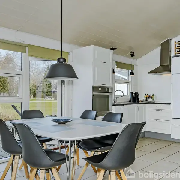 Et moderne køkken-alrum med et hvidt spisebord og sorte stole. Rummet har store vinduer med udsigt til en have. Køkkenet har hvide skabe, køleskab og ovn.
