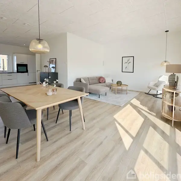 Spisestue med træbord og stole, lys opholdsstue med sofa og tæppe, moderne køkken i baggrunden. Lyst trægulv og minimalistisk indretning med lamper og billeder på væggen.