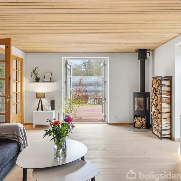 En lys stue med en grå sofa, et ovalt sofabord med en vase blomster, en brændeovn med stablet brænde og en skænk med TV. Dør åbner til terrasse.
