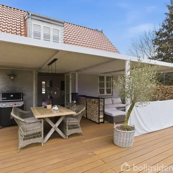 Terrassen har et trædæk og overdækning, møbleret med et bord og stole til afslapning. Grill og loungeområde med sofa og planter skaber hygge. Husets facade er synlig i baggrunden.