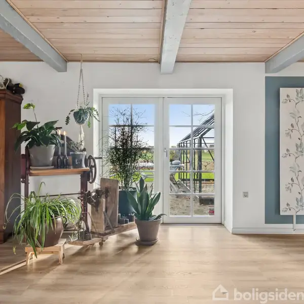 Træmøbler og planter står i en lys stue foran store glasdøre. Udenfor ses en terrasse og have. Væggene har dekorative paneler, og loftet har træbjælker.