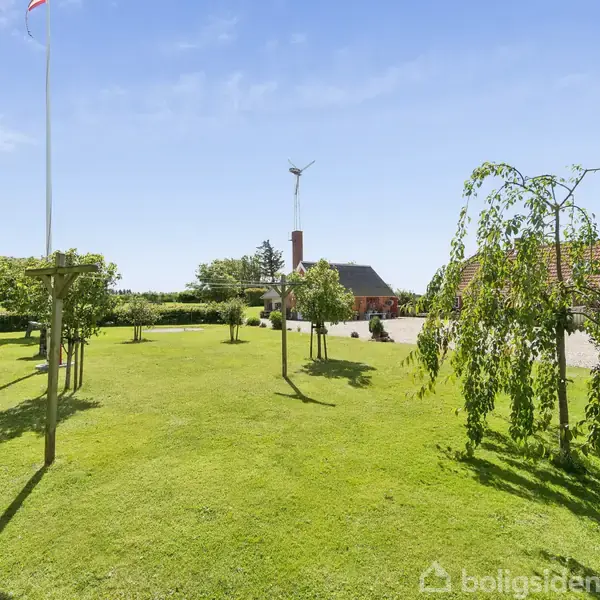 En velplejet græsplæne med spredte træer står foran en gård med rødt tag. En flagstang og en vindmølle ses i baggrunden under en klar, blå himmel.