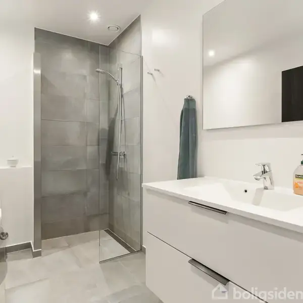 Badeværelse med brusekabine og flisebelagt væg på højre side. Håndvask til venstre med skabsspejl, sæbedispenser og håndklæde. Toilet ved siden af med dekorative genstande ovenpå.