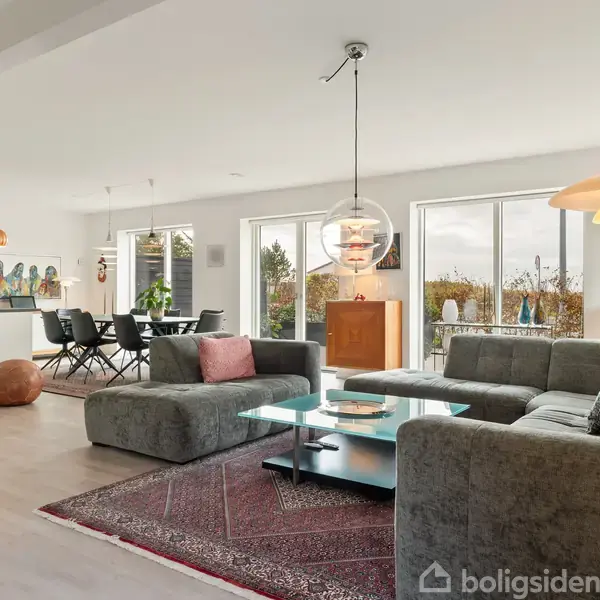 Stue med grå sofaer og glasbord på et tæppe. Store vinduer med udsigt til haven. I baggrunden er der et spisebord og køkken med moderne lamper.