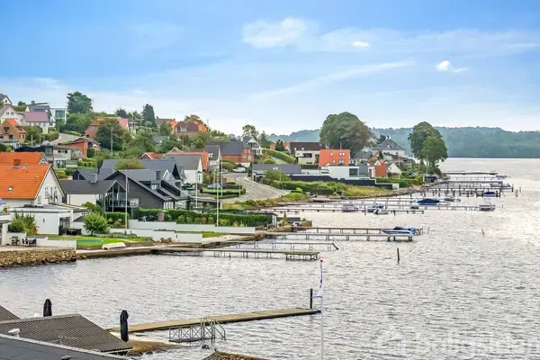 Spisebord med stole står i et rum med røde vægge og trægulv. En skænk med glaslåger er til venstre, og en gulvlampe samt hængende billeder udgør dekorationen.