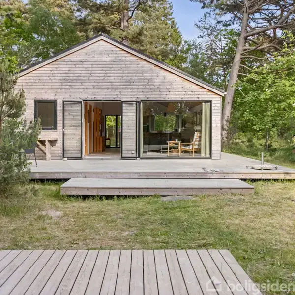Et moderne træhus står på en hævet terrasse omgivet af skovklædte områder. Store glasdøre giver udsigt til en minimalistisk indretning.