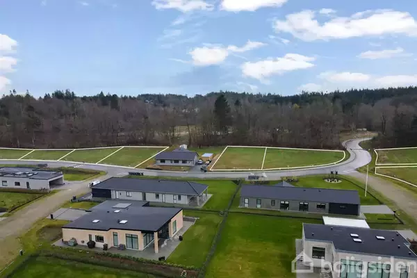Flere moderne huse med flade tage står stille i et boligområde omgivet af velholdte græsplæner og veje, bagved ligger en skov, og himlen er delvist overskyet.