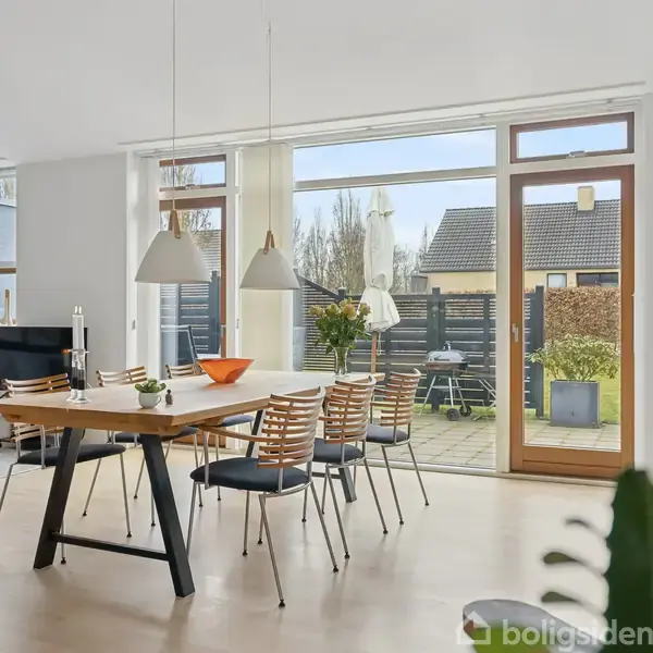 Spisebord omgivet af stole står i et lyst rum med store vinduer og udsigt til en terrasse med grill. Rummet indeholder også en sofa og TV i baggrunden.