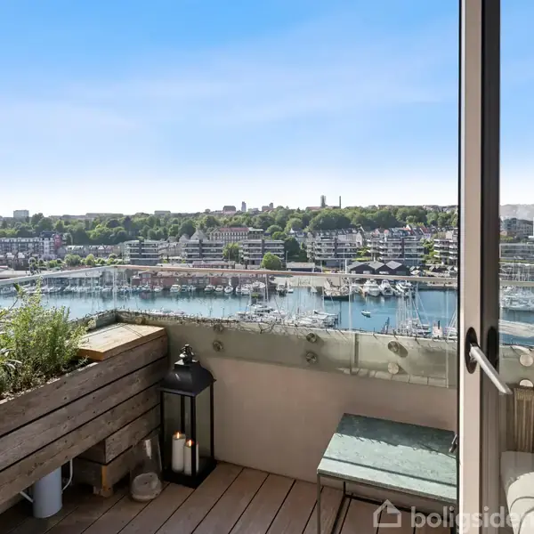 Terrassen er udstyret med en bænk og planter, vender ud mod en marina fyldt med både. Byens skyline er synlig i baggrunden under en klar blå himmel.