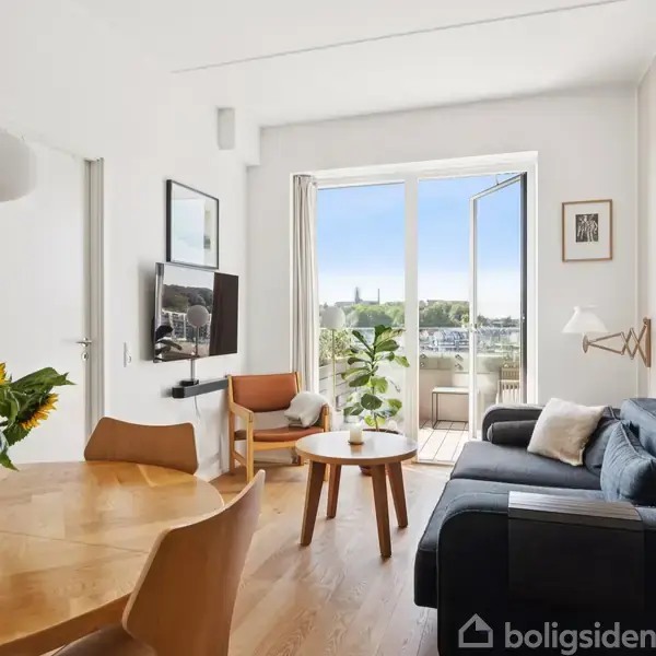 Stue med træbord, solsikker og stole, sofa nær glasdør åben mod balkon, udsigt til bylandskab; moderne indretning, lyse vægge, fladskærms-TV på væggen, dekorative kunstværker omkring.