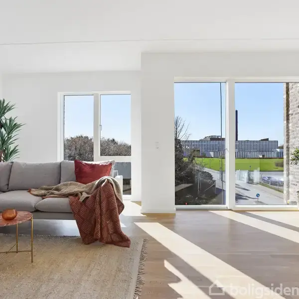En grå sofa med farverige puder står i et lyst stueområde. En stor vinduesfront giver udsigt til en grøn have. Et tæppe og små borde komplementerer rummet.