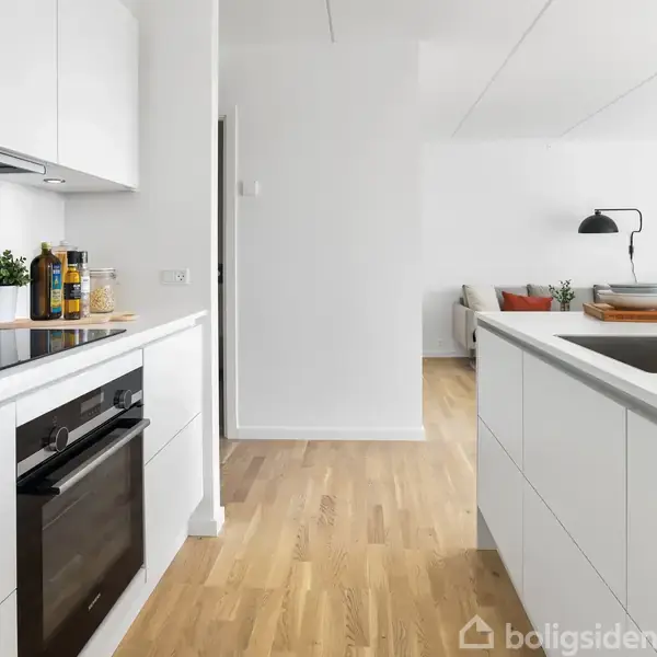 Et moderne køkken med hvide skabe og en sort ovn. En køkkenø med indbygget vask står centralt. I baggrunden ses stueindretning med sofa og planter.