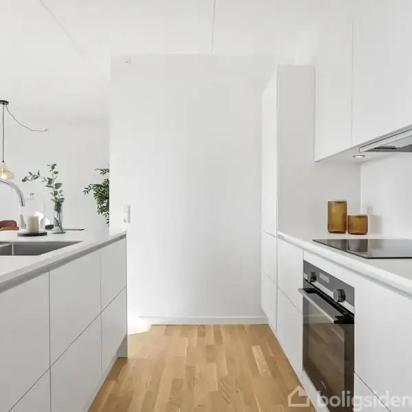 Moderne køkken med hvide skabe og træplankegulv. Køkkenbordet har indbygget vask og ovn, med krydderier og olier på bordet. I baggrunden ses et spiseområde med planter.