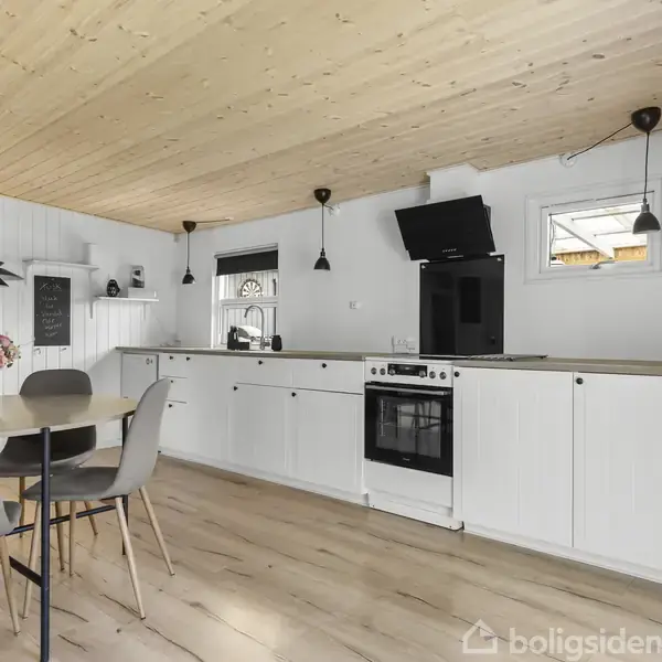 Køkken med hvide skabe, sort ovn og komfur, sort emhætte; rundt spisebord med grå stole. Trægulv og -loft, hvide vægge. Blomster på bordet, redskaber på en skænk.