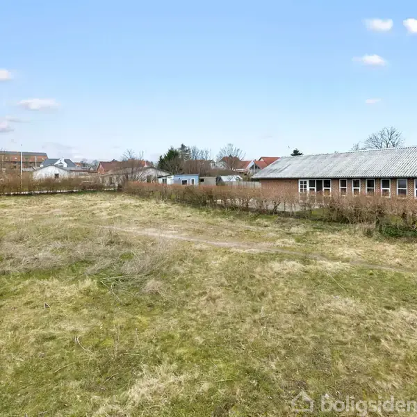 Et åbent græsareal ligger stille ved siden af en vej. I baggrunden ses et rækkehus med flere vinduer. Omkring er et beboelsesområde med huse og haver.