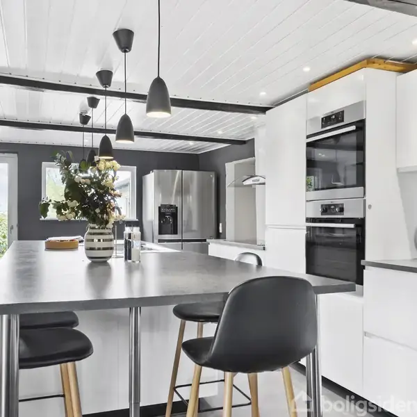 Moderne køkken med en stor grå bordplade omgivet af sorte stole. Hængende lamper oplyser rummet, mens hvide skabe og en grå væg skaber en stilfuld atmosfære. En dør fører udenfor.