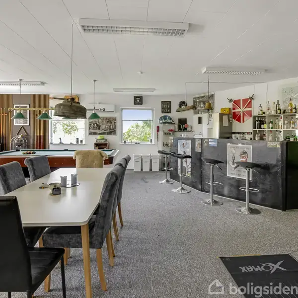 Et rummeligt værelse indrettet som bar og opholdsområde; det har et billardbord, en lang spisebord med stole, barstole foran en bar, og hylder med flasker og dekorationer på væggene.