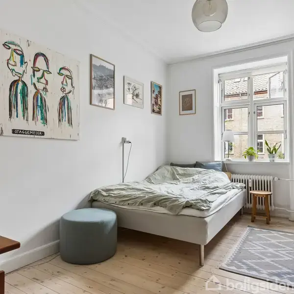 Soveværelse med en seng langs væggen, omgivet af kunstplakater. Et stort vindue tilgår naturligt lys. Skab og skrivebord kompletterer det minimalistiske rum.