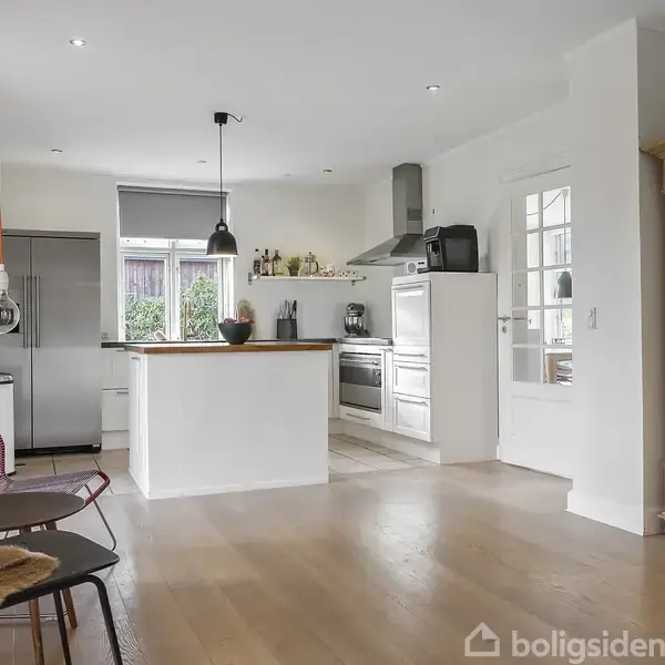 Køkken med hvid indretning, komfur og emhætte midt i rummet, omgivet af træmøbler. En kogeø med hængende lampe, stole med lammeskind i forgrunden, store vinduer i baggrunden.