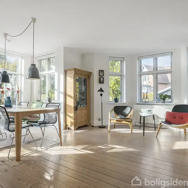 Et lyst spisestueområde med et træbord og seks stole, omgivet af store vinduer. I hjørnet står en vitrineskab, og forskellige moderne lænestole i en solrig atmosfære.