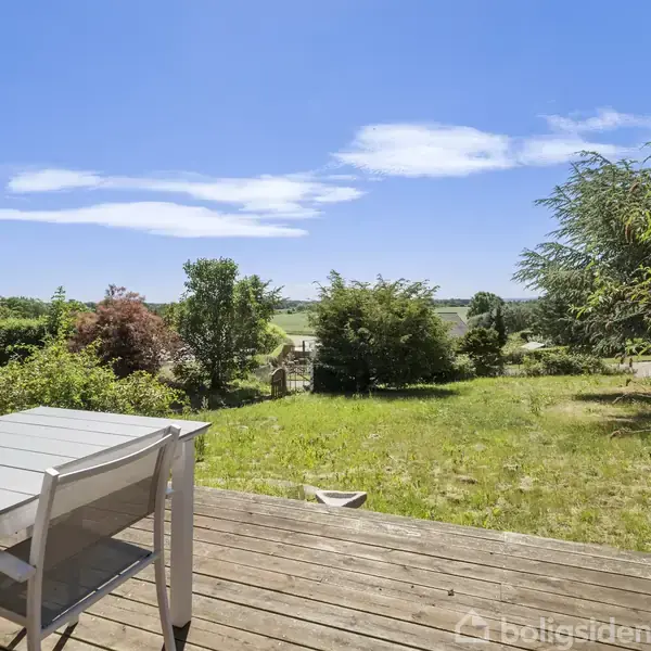Terrassebord og stole står på en træterrasse. I baggrunden ses en stor have med græs og buske omkranset af træer. Himlen er klar blå med få skyer.