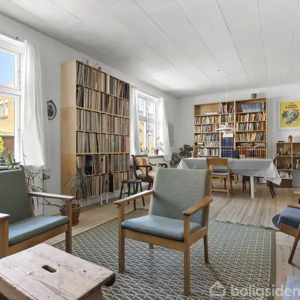 Stue med træmøbler og grønne lænestole arrangeret omkring et lavt bord, med bogreoler fyldt med bøger i baggrunden, to store vinduer med hvidt gardin, og en spiseplads ved et dækket bord.