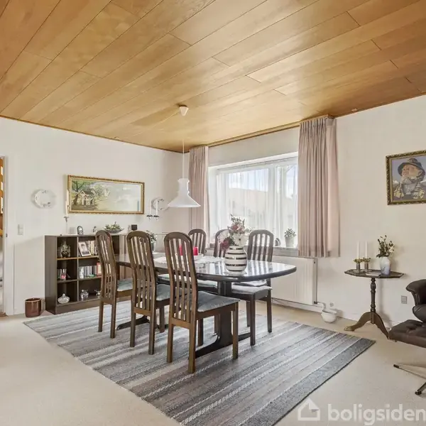 Spisestue med træbord og seks stole under en loftslampe. Stueplan har træloft, tæppe, reol, lænestol og store vinduer med gardiner, som skaber en lys og varm atmosfære.