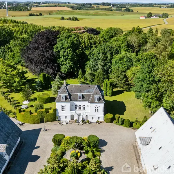 En hvid, herregårdslignende bygning står mellem to lader og omgivet af frodige træer. Omkring bygningen er en velholdt have med blomsterbede og en indkørsel. Landskabet er landligt.