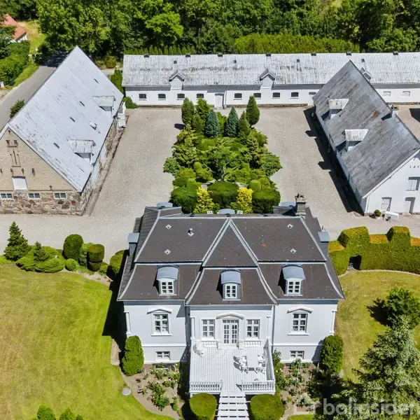 En stor, hvid herregård med sort tag i centrum omkranset af grønne træer og buske. Den ligger foran to store udbygninger med hvide tage, som omkranser en gårdsplads.