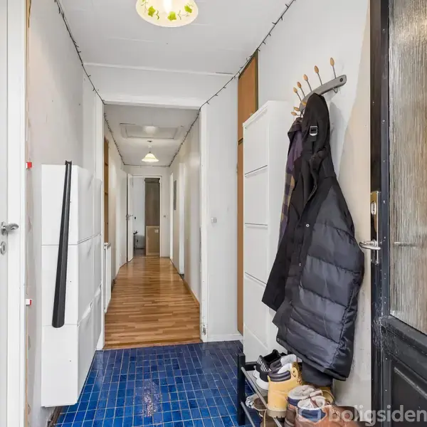 En gang har en jakke hængende på knager, mens sko står på gulvet. Blåt klinkegulv i forgrunden og trægulv i baggrunden, omgivet af hvide vægge og døre.