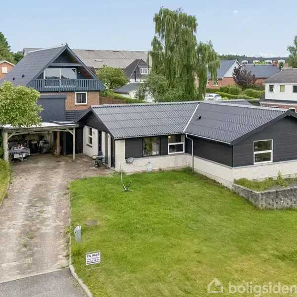 En sort/hvid bungalow ligger på en velplejet græsplæne med en indkørsel og carport til venstre. Omgivet af andre huse i et beboelseskvarter. Skilt: "Til salg" ved vejen.