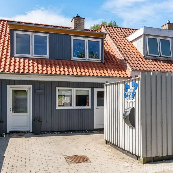 Et rækkehus med gråt træbeklædning og rødt tegltag. Foran huset er en flisebelagt indkørsel. Ved indkørslen er en skraldestation afskærmet af et hegn.