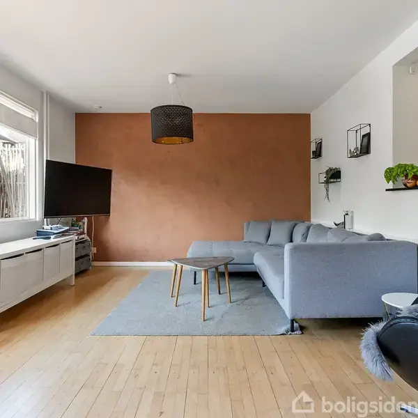 En stue med en grå hjørnesofa placeret mod en brun væg. Et fladskærms-tv står på en hvid medieenhed ved vinduet. Trægulv og moderne indretning præger rummet.