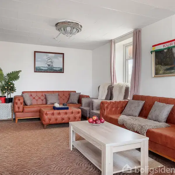 Brune lædersofaer med puder står i en stue. Et sofabord med en frugtskål foran. Rummet har billeder på væggene, inklusiv en med teksten "KURDISTAN".