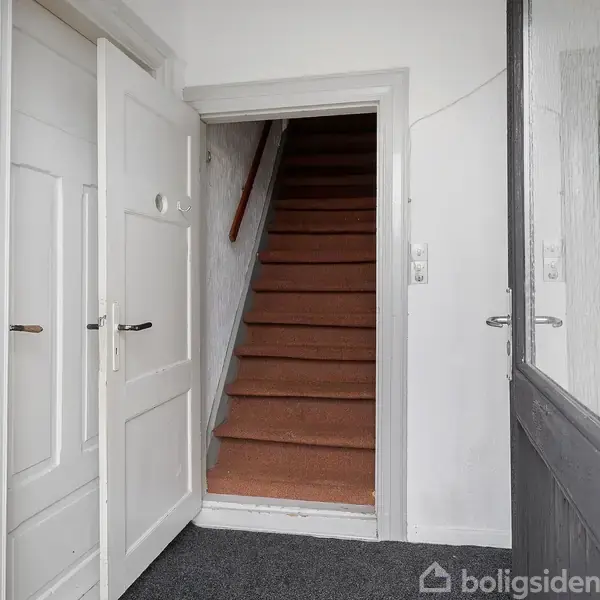 En trappe med brunt tæppe fører opad fra en lille gang. Kontexten inkluderer en åben hvid dør til venstre, og en grå dør til højre.