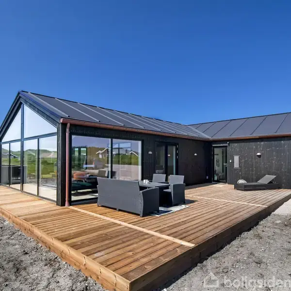 Et moderne, sortmalet sommerhus med store glaspartier og fladt tag. Huset står på en træterrasse med havemøbler. Den klare blå himmel og grønne omgivelser giver en lys og åben atmosfære.