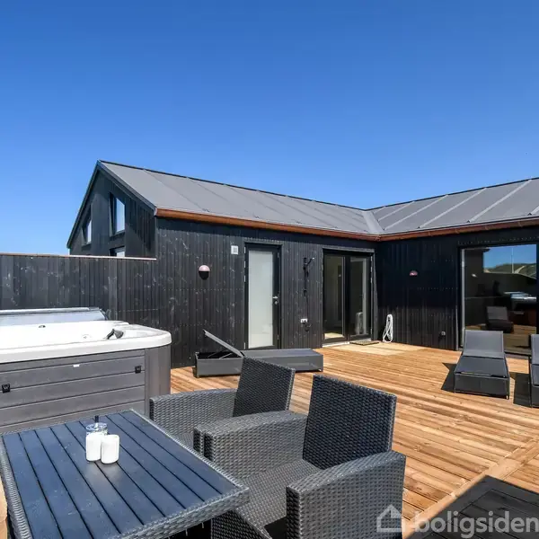 Et udendørs terrasseområde med spabad og loungemøbler. Området er omkranset af et moderne, sort træhus. Blå himmel ses i baggrunden, hvilket indikerer en solrig dag.