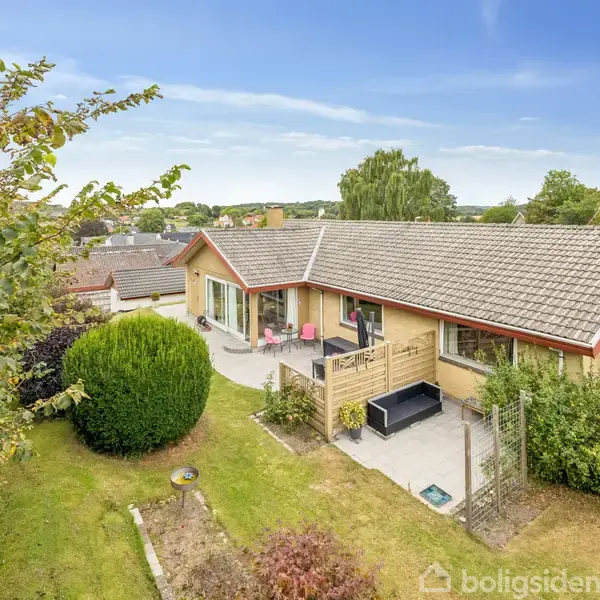 En gul murstensvilla med gråt tag ligger i en have med grønne buske og græsplæne. Terrassen har havemøbler og er omkranset af en lav trævæg.