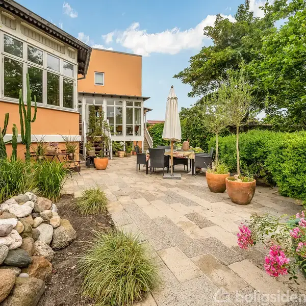 Terrasse med flisebelægning, bord og stole under en parasol. Omkranset af potteplanter, blomsterbede og buske ved siden af et hus med store vinduer.