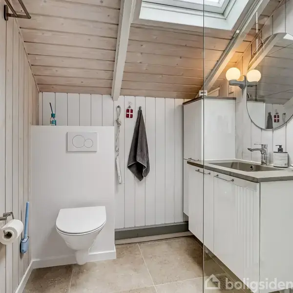 Toilet hænger på væggen, ved siden af hvidt skab med håndvask. Rundt spejl ovenfor skabet. Håndklæde hænger på væggen. Loftsvindue giver lys. Vægge og loft i træ.