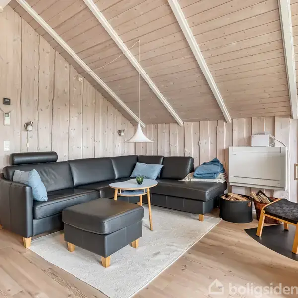 Sort læderhjørnesofa med skammel står i stue. En moderne brændeovn i hjørnet, ild tændt. Trævægge og skråloft skaber en hyggelig atmosfære. Enkelt sofabord foran sofaen.