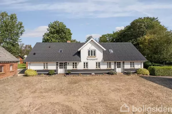 Hus med skifertag og hvid facade, markeret med teksten "Friemannsgaard". Det står på en bar jordgrund, omgivet af træer og buske, placeret i en landlig miljø.