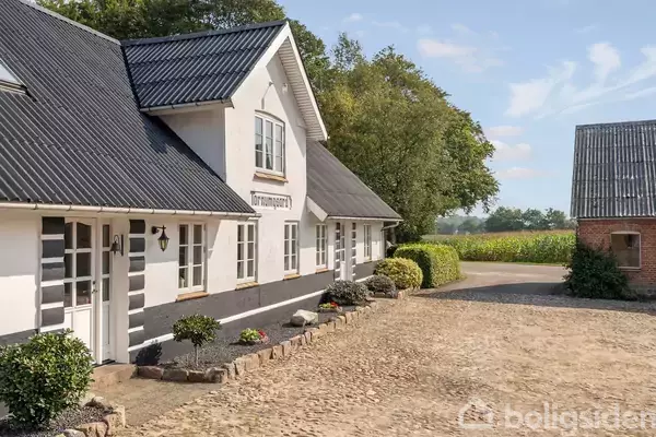 Et hvidt, traditionelt landhus med sorte tagplader står ved siden af en brostensbelagt gårdsplads. Omgivet af buske og træer, ses marker i baggrunden. Skiltet lyder "Smorhjemgård".